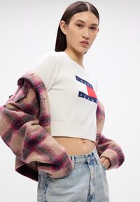 Biely skrátený sveter s námorníckym a červeným logom 'TOMMY JEANS', doplnený o oversized kabát s ružovo-sivou kockou a svetlomodré džínsy.