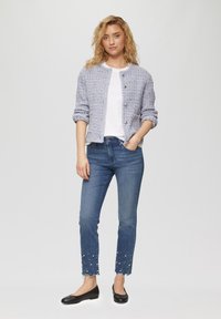 Blauwe tweedjas met ronde hals en zilveren knopen, gecombineerd met slim-fit jeans met een rafelmouw en versierd met witte kralen.