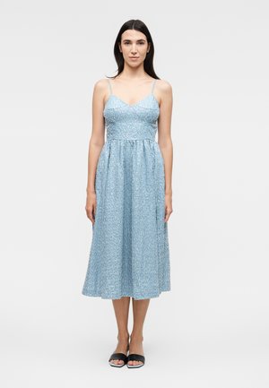 Kobieta stoi, ubrana w bezrękawnikową, jasnoniebieską, wzorzystą sukienkę midi z cienkimi ramiączkami i czarnymi sandałami z odkrytymi palcami na jednolitym białym tle.