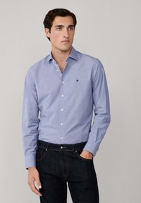 Uomo che indossa una camicia azzurro chiaro a maniche lunghe con un piccolo logo sul petto e jeans blu scuro, in piedi contro uno sfondo chiaro e uniforme.