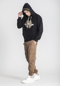 Sudadera negra con capucha y gráfico dorado, combinada con pantalones cargo beige que tienen múltiples bolsillos y zapatillas blancas.