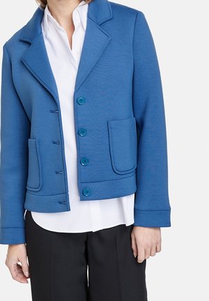 Femme portant un blazer bleu boutonné avec deux poches avant par-dessus une chemise blanche et un pantalon noir sur un fond uni.
