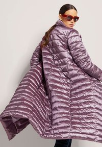 Lange, opbollende jas van metallic lavendel stof met een horizontaal gequilt patroon en een hoge kraag. Heeft een vloeiende silhouet en zijsplitten.