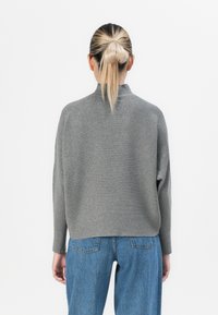 Person with blonde hair in a low bun wearing a gray ribbed sweater and blue jeans, standing with back facing the viewer against white background.