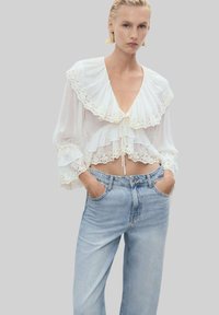 Vit blus med en rynkad spetskrage och lagerdesign, tillsammans med högmidjade ljusblå jeans. Blusen har dragsko och urringade ärmar.