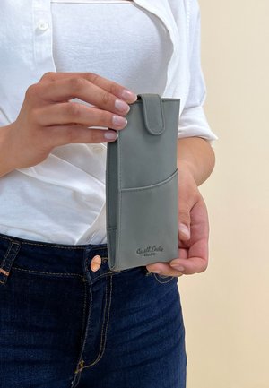 Šedý kožený kryt na telefon s kapsou, s uzávěrem na knoflík a výrazně embosovaným logem značky. Držen v ruce na světlém pozadí.