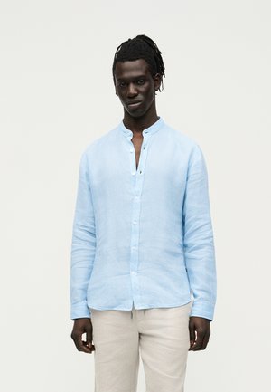 Young man with dark skin and dreadlocks wearing a light blue button-up shirt and beige pants, standing against a plain light background.