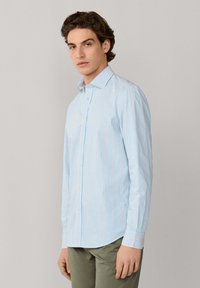 Jeune homme portant une chemise à manches longues bleu clair et blanc à rayures et un pantalon vert olive, debout devant un fond uni.