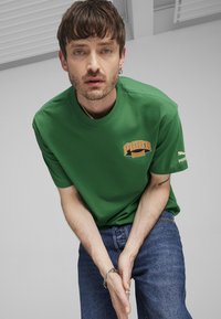 Camiseta de algodón verde de cuello redondo con el logo naranja "Puma Sports", de manga corta, combinada con pantalones vaqueros azules. Diseño simple y casual.
