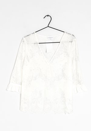 Hvid bluse i blonder med V-hals, 3/4-ærmer med flæser og bølget kant, hængende på en sort tråd-bøjle mod en hvid baggrund.