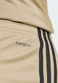 Béžové sportovní šortky s černými pruhy po stranách, vyrobené z texturované látky a s černým brandingem "AEROREADY" poblíž pasu.