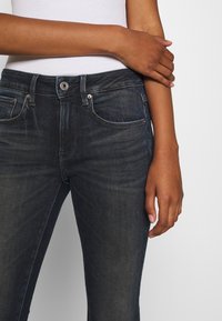 Osoba v tmavě modrých džínách a bílém topu, s jednou rukou opřenou o protilehlou ruku před jednobarevným pozadím.