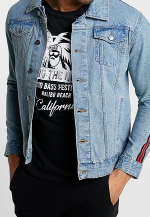 Homme portant une veste en jean bleu clair par-dessus un t-shirt noir à motif Malibu Beach California, les mains dans les poches.