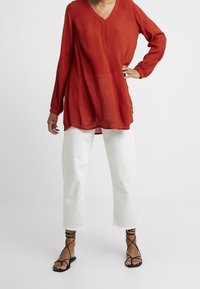 Haut rouge ample à manches longues avec col en V, associé à un jean blanc court et des sandales noires à brides. Tissu texturé.