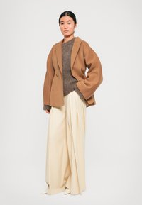 Veste double boutonnage en mélange de laine marron, pull en tricot gris, pantalon large crème et escarpins pointus beiges. Textures lisses, design minimal.