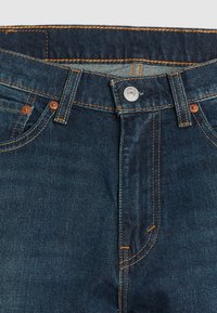 Donkerblauwe denim jeans met koperen klinknagels, oranje stiksels, een metalen knoopsluiting en vijf zakken. Gladde textuur met een klassieke snit.