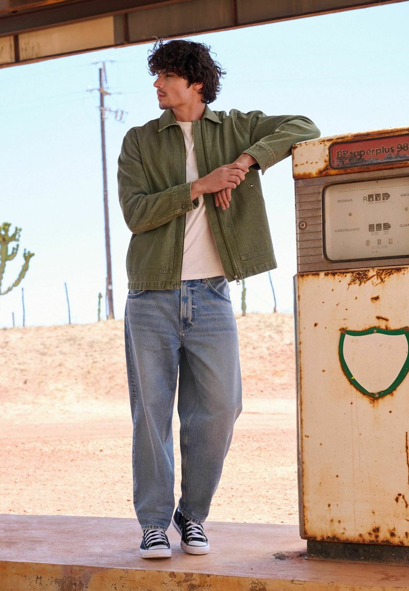 Joven con chaqueta verde y vaqueros apoyado en una antigua surtidora de gasolina oxidada en una estación de carretera en el desierto.