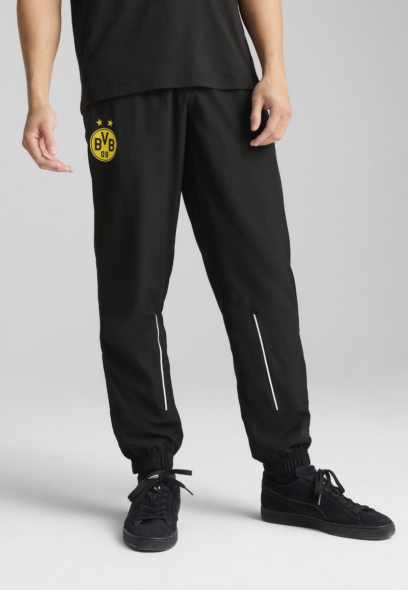 Pantalones deportivos negros con un logotipo circular amarillo que dice "BVB 09", rayas reflectantes blancas y puños elásticos. Fabricados con un material ligero.