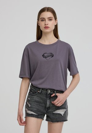 T-shirt grigio a maniche corte con grafica di un cuore e testo, abbinato a pantaloni corti di denim nero sfilacciati con orli sfrangiati e usura visibile.