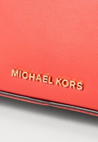 Sac en simili cuir corail avec un logo "MICHAEL KORS" en relief doré. Texture lisse avec des accents noirs le long des bords.
