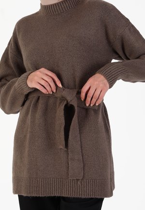 Personne attachant une ceinture en tricot marron de pull à la taille, montrant des poignets et un ourlet côtelés, vêtue d'un pantalon noir sur un fond blanc.