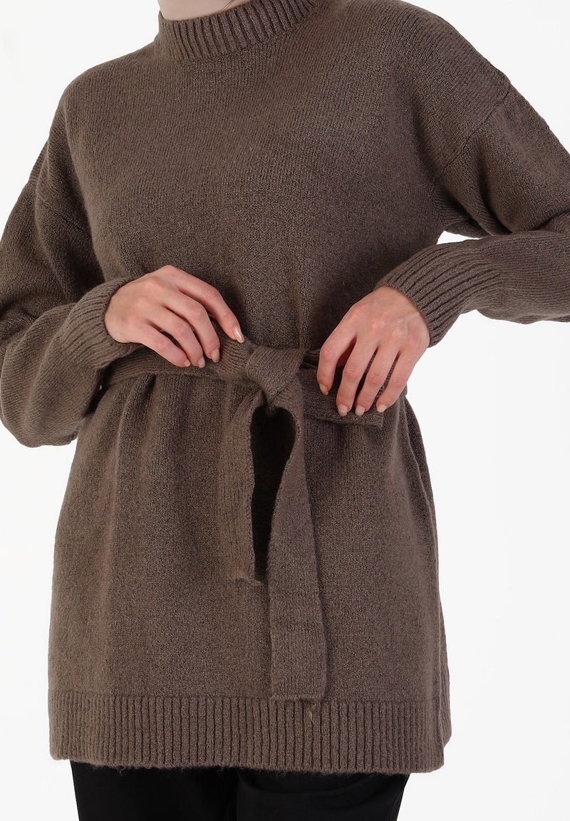 Personne attachant une ceinture en tricot marron de pull à la taille, montrant des poignets et un ourlet côtelés, vêtue d'un pantalon noir sur un fond blanc.