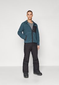 Chaqueta de forro polar en color verde azulado con detalles en negro, con cremallera frontal y bolsillos laterales, combinada con pantalones negros y botas. Textura suave.