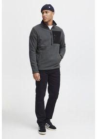 Pullover grigio in pile con una tasca nera con zip; colletto con zip; indossato con jeans neri e scarpe nere con tre strisce bianche.