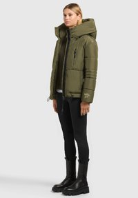 Doudoune olive avec un col montant et une grande capuche, dotée de poches zippées et d'une finition texturée, portée par-dessus un legging noir et des bottes.