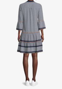 Femme de dos, portant une robe imprimée longueur genoux avec des manches évasées et des baskets blanches sur fond blanc.