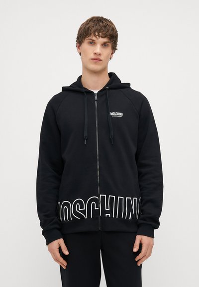 Zwarte zip-up hoodie gemaakt van zachte stof, met een logo op de borst en grote witte letters aan de onderrand. Met een aantrekkoord in de capuchon en raglan mouwen.