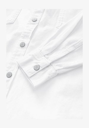 Valge nööpideta särk, millel on hõbedased trukk-nööbid, lame krae ja pikad varrukad koos kokku volditud mansettidega, mis näitavad siledat ja tekstuurset kangast.