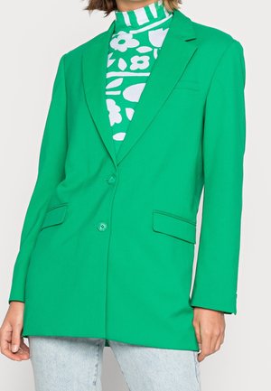 Mujer con blazer entallado de color verde brillante con dos botones sobre un jersey de cuello alto con estampado floral en verde y blanco, combinado con vaqueros azul claro.