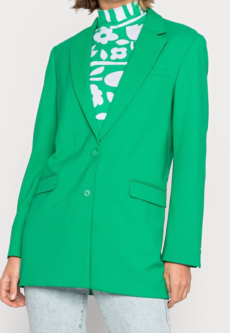 Femme portant un blazer vert vif ajusté à deux boutons sur un col roulé à motifs floraux verts et blancs, associé à un jean bleu clair.