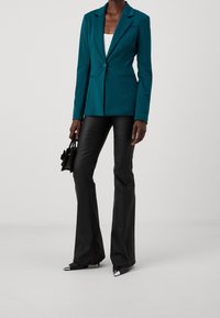 Kvinna i tealfärgad blazer, vit topp, svarta utsvängda byxor, svarta spetsiga skor med metalltå, hållande en liten svart handväska.