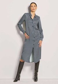 Knielanges Hemdblusenkleid in Marineblau mit weißem kreisförmigem Muster, langen Ärmeln und einem hohen Schlitz. Kombiniert mit knielangen schwarzen Stiefeln. Glatter Stoff.