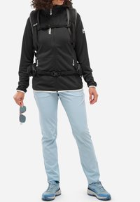 Veste polaire zippée noire avec des accents gris, pantalon de sport bleu clair, et chaussures de randonnée bleues. Deux sacs à dos, lunettes de soleil tenues à la main.