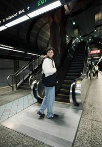 Scala mobile in una stazione della metropolitana con un pavimento grigio texturizzato, un segnale di uscita verde sopra e una persona in giacca crema e jeans blu che si trova nei pressi.