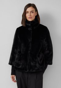 Veste en faux fourrure noire avec un col montant, des �épaules tombantes et deux poches avant. Sa texture douce et sa forme trapèze accentuent la chaleur.