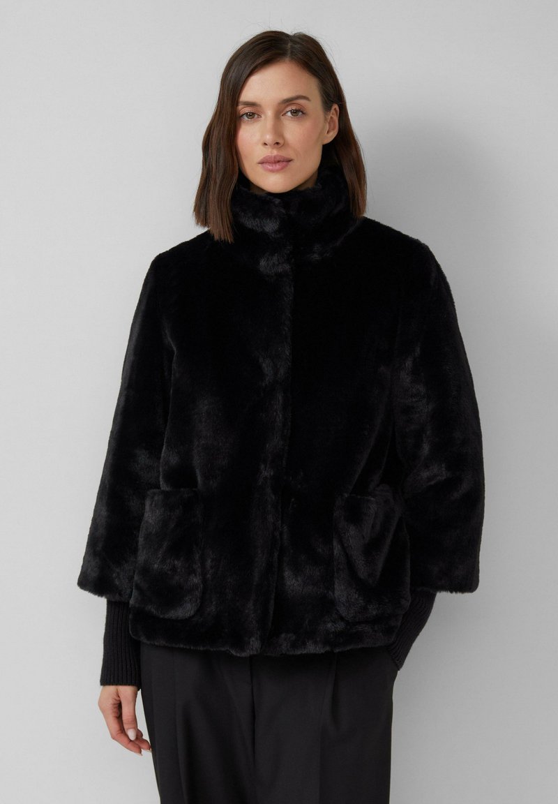 Veste en faux fourrure noire avec un col montant, des épaules tombantes et deux poches avant. Sa texture douce et sa forme trapèze accentuent la chaleur.