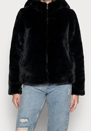 Person iført en sort faux fur jakke med lynlås og lyseblå hullede jeans, stående med fronten frem mod en ensfarvet lys baggrund.