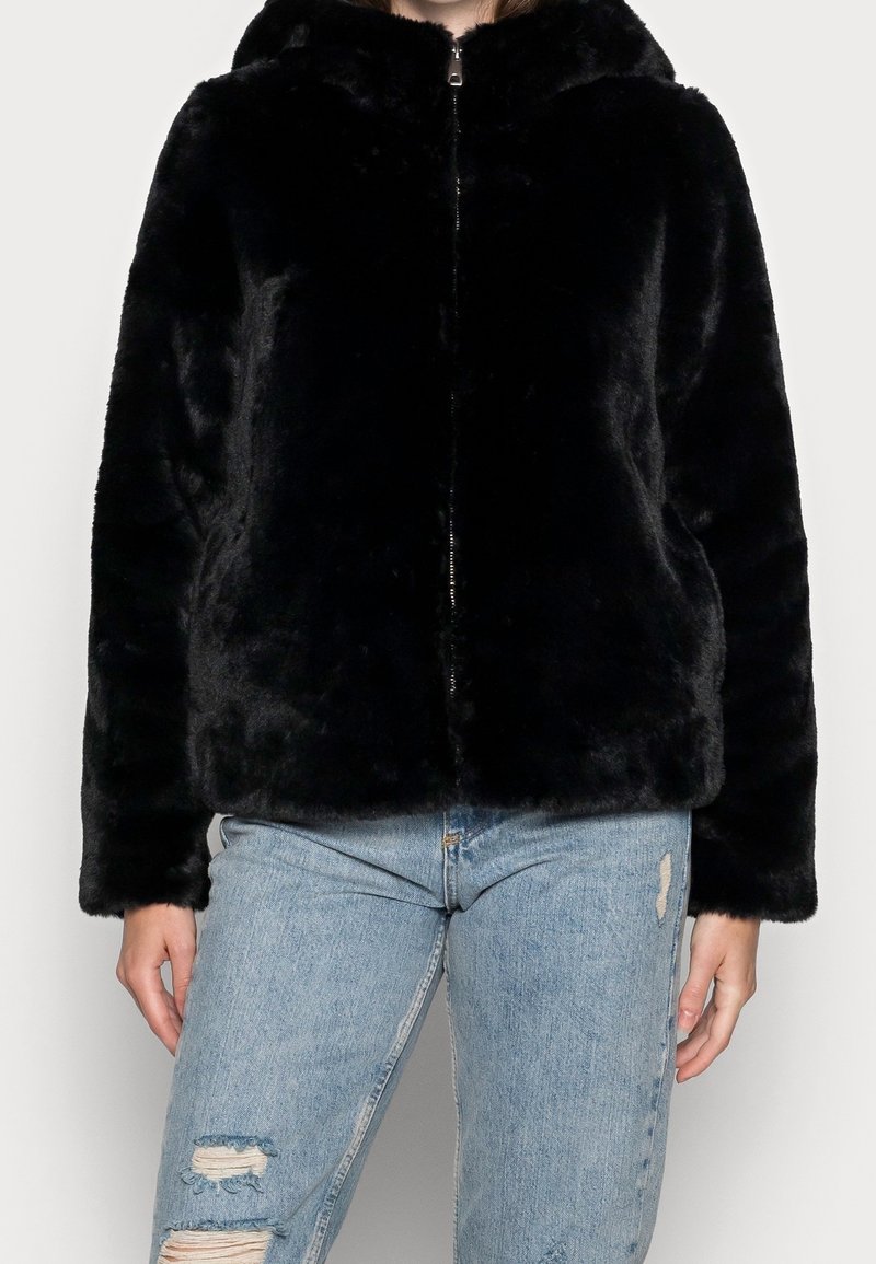 Personne portant une veste noire en fausse fourrure à fermeture éclair et un jean bleu clair déchiré, de face sur un fond clair et uni.
