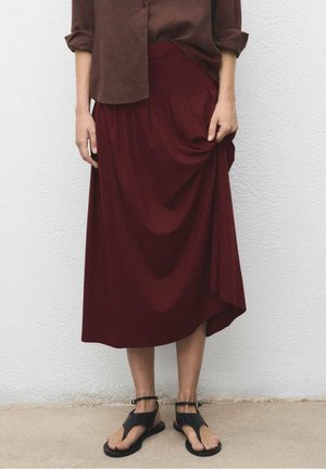 Personne portant une jupe midi bordeaux, une chemise marron à manches longues et des sandales noires à bout ouvert, debout contre un mur gris clair.