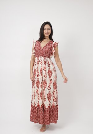 Mujer de pie con un vestido maxi sin mangas de patrón en crema y rojo óxido, con detalles de volantes en los hombros, sonriendo frente a un fondo liso.