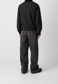 Person klädd i svart jacka och löst sittande mörkgrå jeans står mot en enkel ljusgrå bakgrund, vänd bort.
