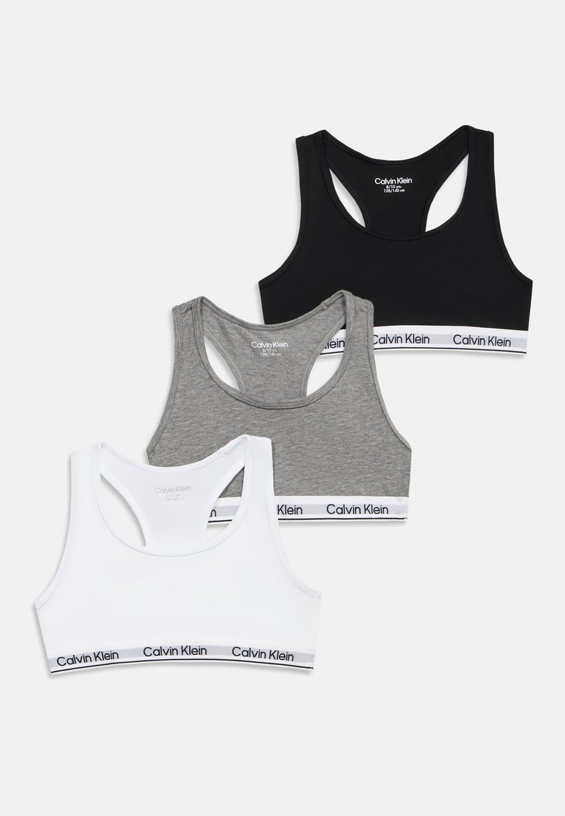 Τρία αθλητικά σουτιέν Calvin Klein racerback σε μαύρο, γκρι και λευκό χρώμα με λωρίδες λογότυπου, τοποθετημένα σε απλό ανοιχτό φόντο.