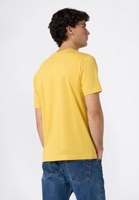 Mann mit lockigem dunklem Haar, der von hinten schaut, trägt ein einfaches gelbes Kurzarmhemd und blaue Jeans mit den Händen in den Taschen.