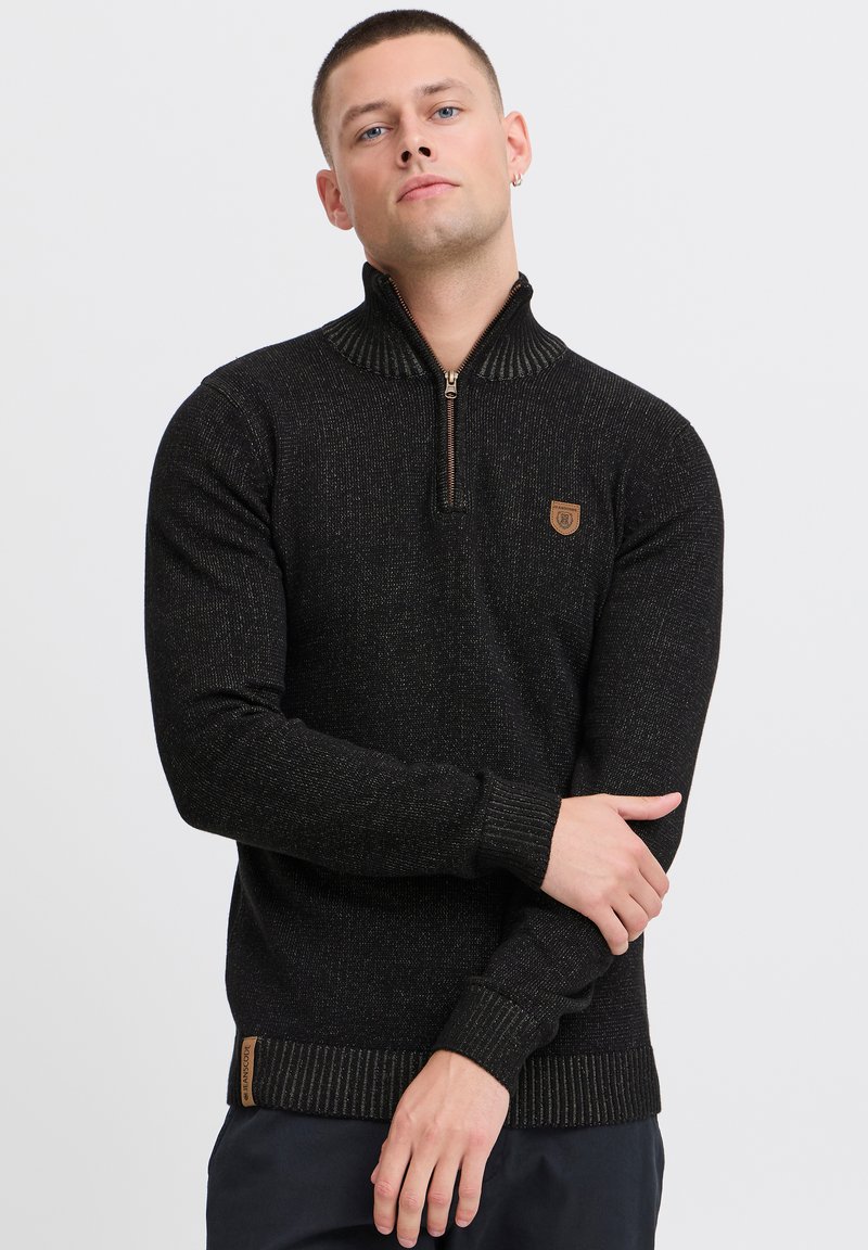 Schwarzer Reißverschluss-Pullover mit geripptem Kragen, der subtile metallische Fäden enthält. Mit einem kleinen braunen Logo-Patch auf der Brust und gerippten Bündchen.