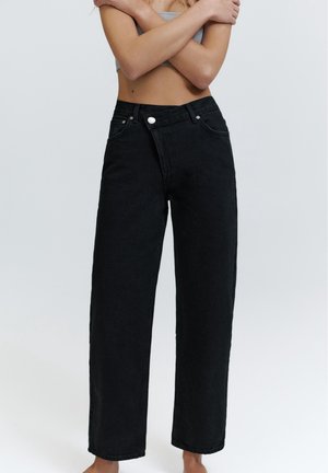 Vrouw draagt loszittende zwarte jeans met hoge taille en een witte knoop, armen gekruist over blote buik tegen een lichtgrijze achtergrond.