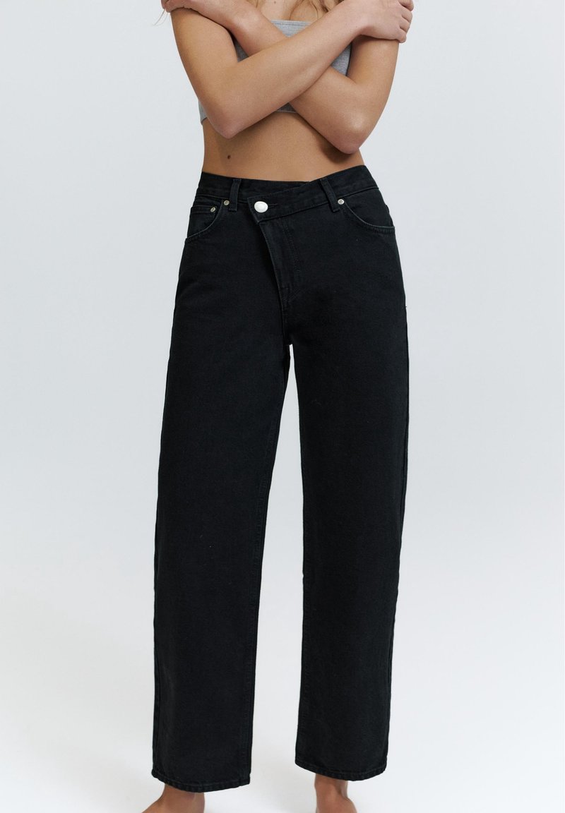Femme portant un jean noir ample taille haute avec un bouton blanc, les bras croisés sur le ventre nu sur fond gris clair.